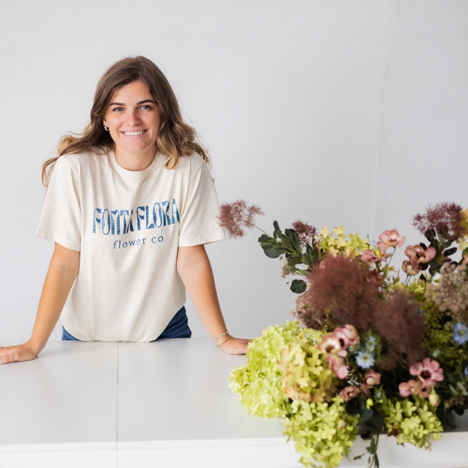Woman standing behind a counter with floral arrangements, wearing a 'Fonta Flora' t-shirt.