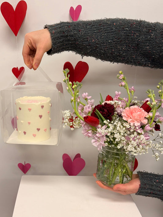 Person holding a bouquet of flowers next to a small cake with heart decorations, against a wall with heart-shaped decorations.